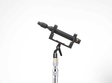 This is a photo of the CO-100K Sanken microphone. The microphone is black and on a side facing view. Held up with a seperate piece of equipment that is black and silver in colour. It is on a white background.